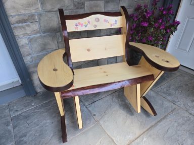 Wooden rocking chair with hand painted decorative flowers on the backrest and armrests, set against a stone wall and flowers.
