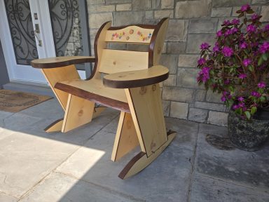 Wooden rocking chair with decorative backrest, placed on a stone patio near flowers.